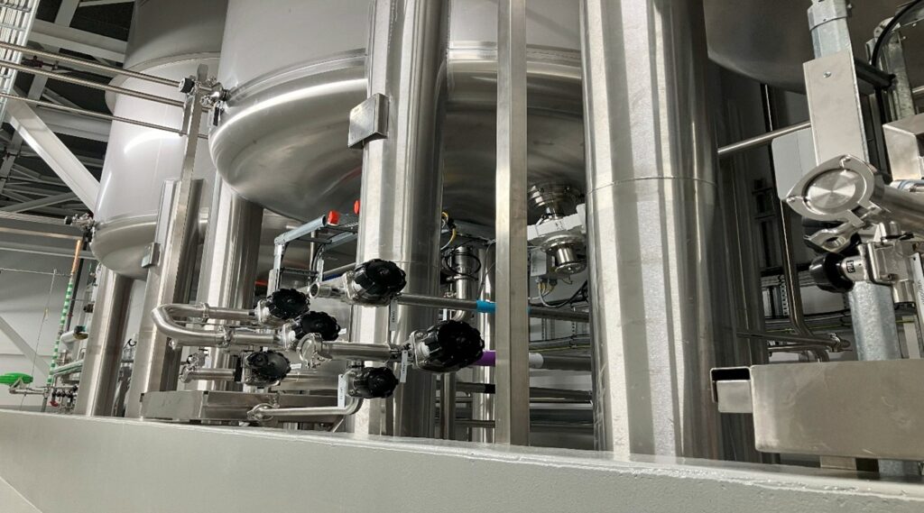 Stainless steel pharmaceutical feed tank integrated in a process system with surrounding pipes and valves for hygienic fluid management.